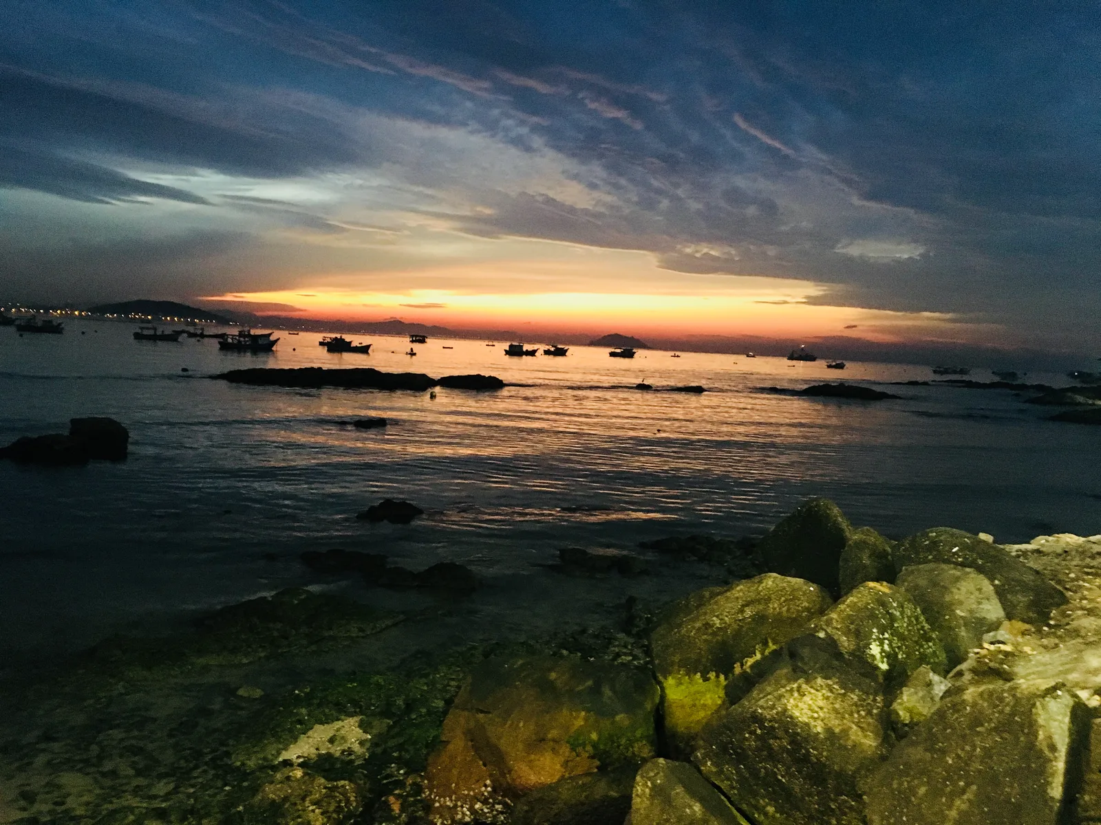 Barcos e pedras no Trapiche de Penha, com céu alaranjado ao pôr do sol